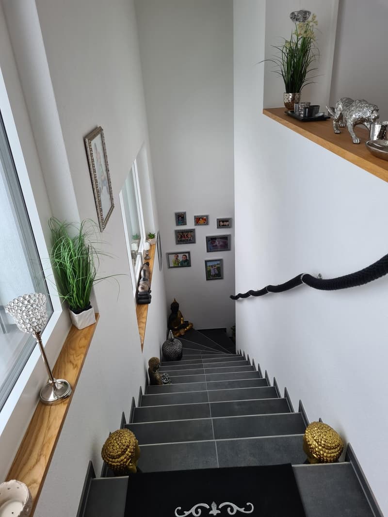 A staircase with multiple steps. White walls and gray steps. A wooden window ledge on the left side with a potted plant and a decorative lamp.