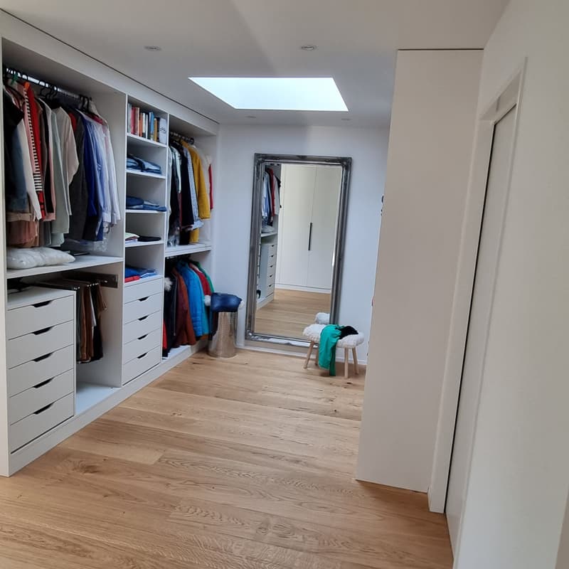 Large walk-in wardrobe with various compartments and drawers. Mirror with frame on wall and trash can. Wooden floor.
