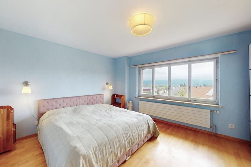 a cozy bedroom with blue walls, wooden floors, a large bed with a white quilt, bedside lamps, a nightstand, and a large window with a view