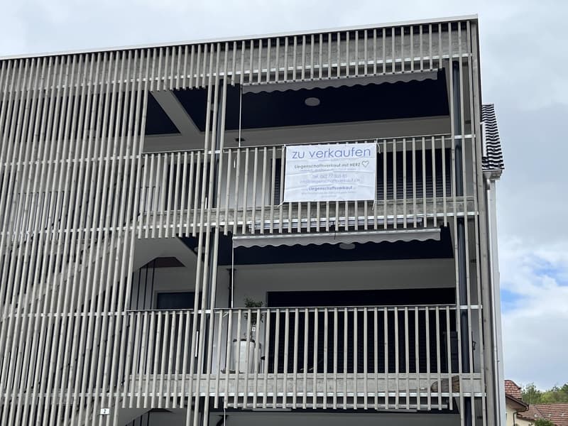 Modern apartment building with balconies, metal railings, and a sign indicating it is for sale
