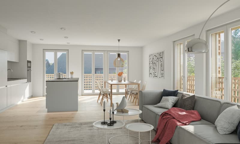 The image shows an open concept living room and kitchen area with large windows providing ample natural light. The kitchen features a modern, minimalist design with white cabinets and a central island. The living room has a gray sofa and a dining table wi