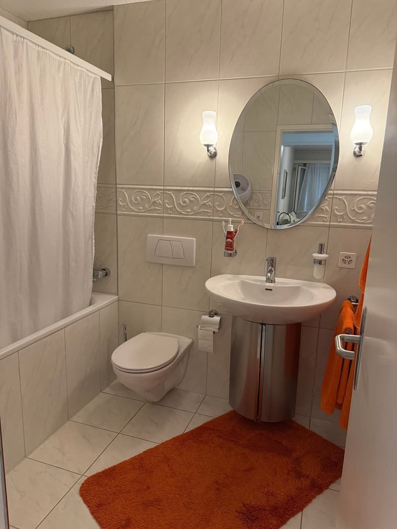 white tiles, round mirror, sink with silver stand, toilet, bathtub, white shower curtain, orange towel
