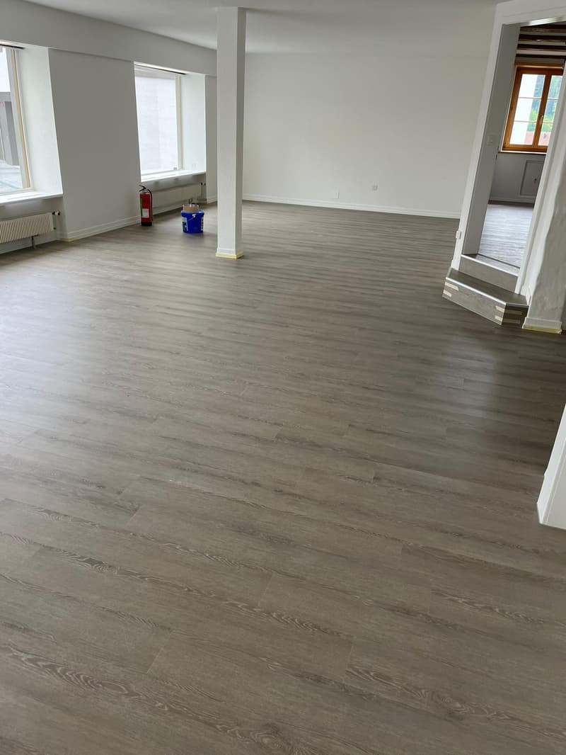 Wide open room with hardwood floors, white walls, and windows. A fire extinguisher is leaned against a wall.