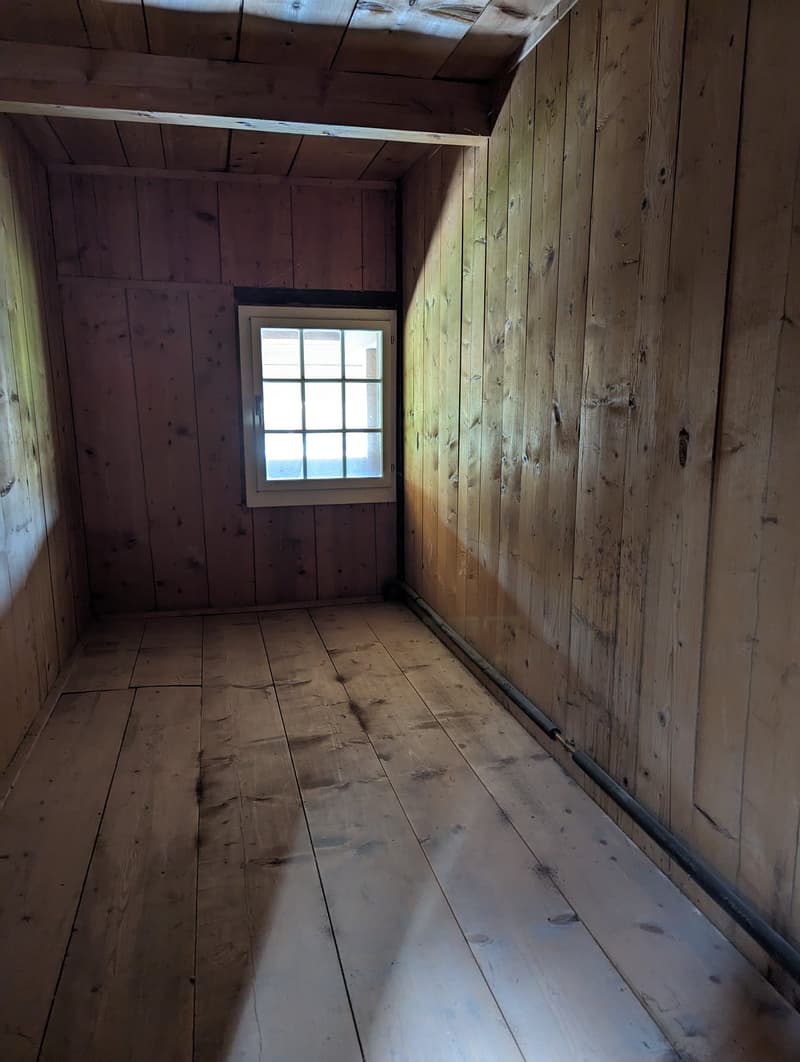 This image shows the interior of a small, rustic room. The walls are made of weathered, wooden planks, and the floor is also wooden. There is a single window with a grid pattern, providing natural light to the space. The room appears to be in a state of d