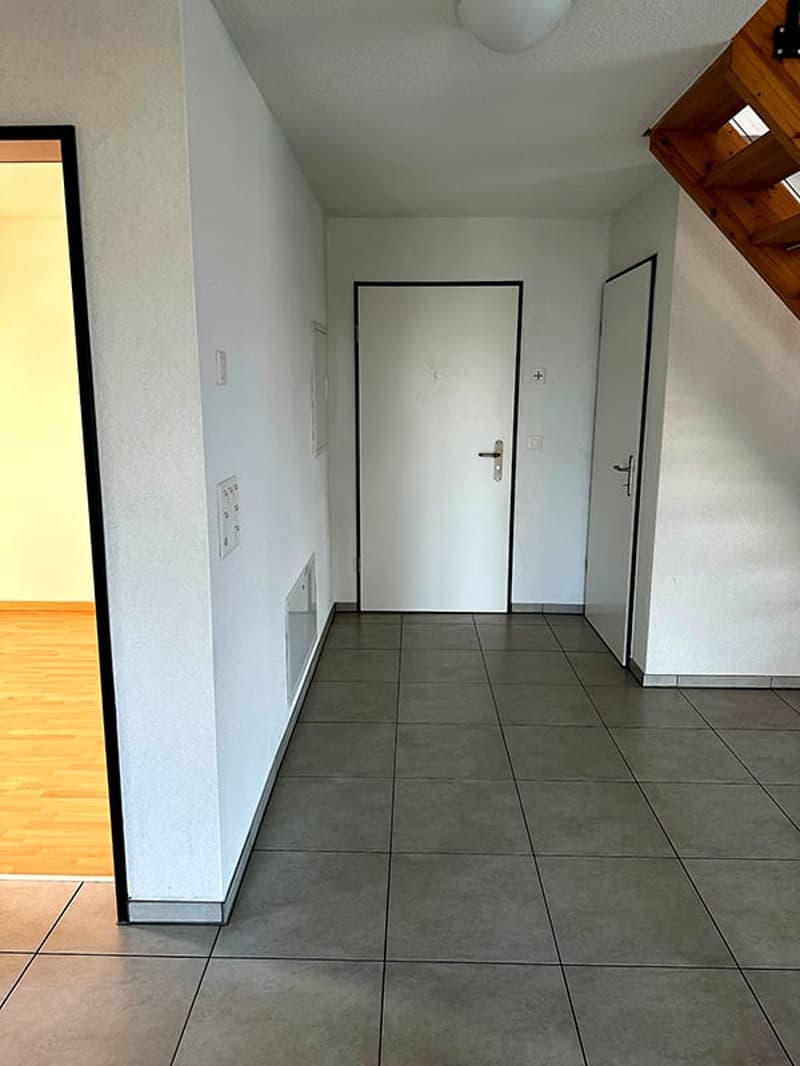 Empty hallway with white walls, gray tiles, two doors, and a wooden staircase.