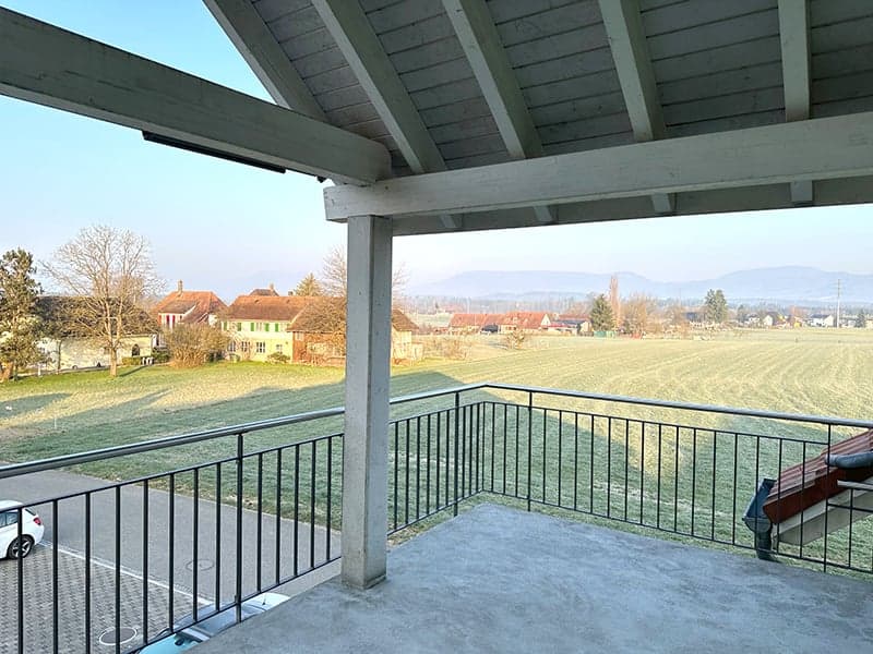 covered balcony with metal railings, overlooking a field and houses