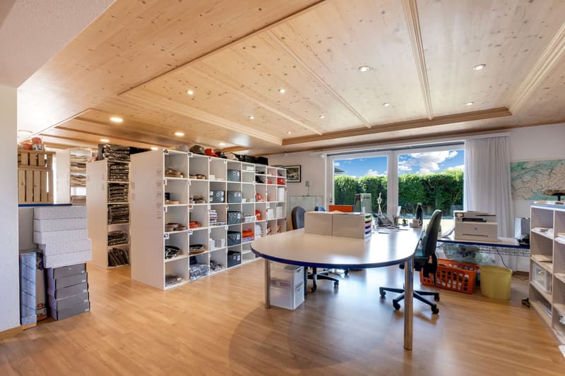 Spacious home office with wooden ceiling, recessed lighting, large windows overlooking the garden, a round table with chairs, and extensive shelving units for storage and organization.