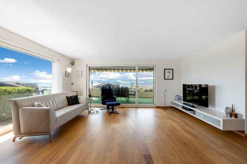 Spacious living room with large windows offering a scenic view, hardwood flooring, beige sofa, TV stand, and a balcony or terrace area