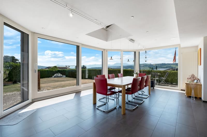 Spacious dining room with large windows offering a scenic view of the surrounding landscape. The room features a wooden dining table with red leather chairs, and the floor is covered with dark tiles. The room has a modern and open design, allowing natural