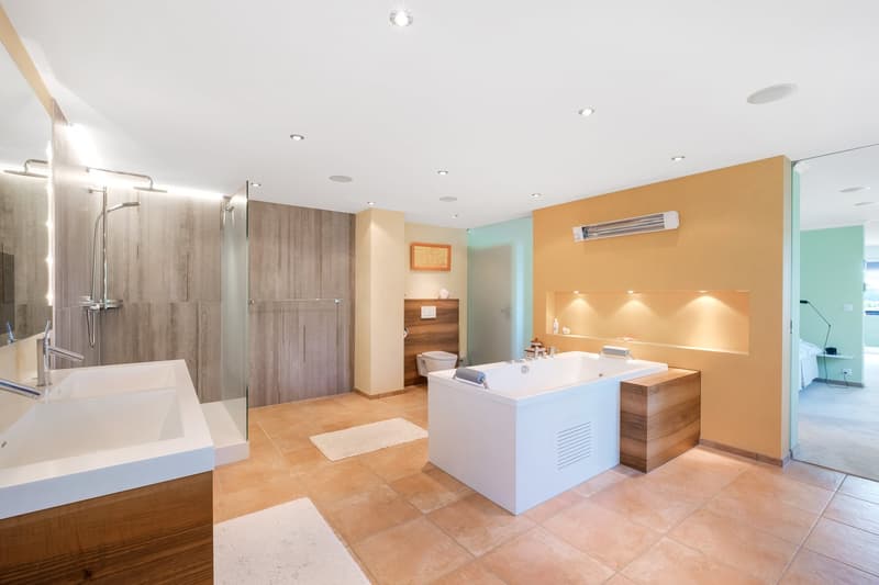 Spacious bathroom with a large bathtub, separate shower area, vanity unit, and built-in storage cabinets. The walls are a combination of neutral tones and warm yellow accents, creating a modern and relaxing atmosphere. The room is well-lit with recessed l