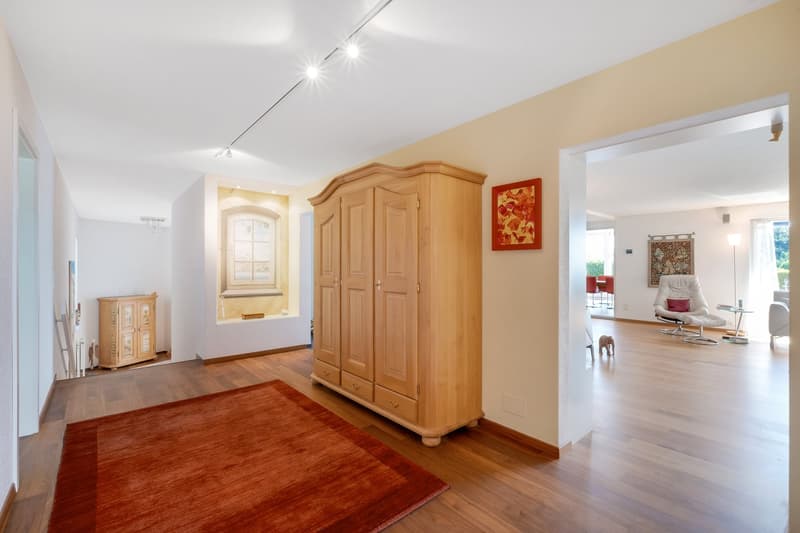 Spacious living room with hardwood floors, large wooden armoire, red area rug, and artwork on the walls. The room has recessed lighting and appears to be connected to other rooms in the home.