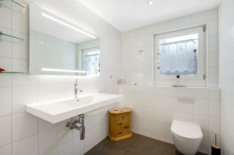 white tiles, white sink, wooden cabinet, white toilet, mirror, window with curtains