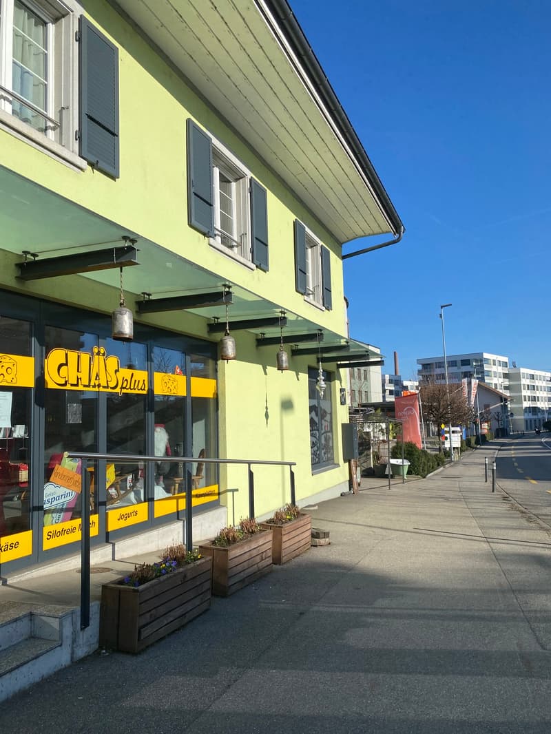 A yellow building with glass windows and doors, a green awning, and a sign that reads 'CHAI's plus'. There are three wooden planters with plants in front of the building. The building has a balcony with a railing and a lamp hanging from the ceiling. There