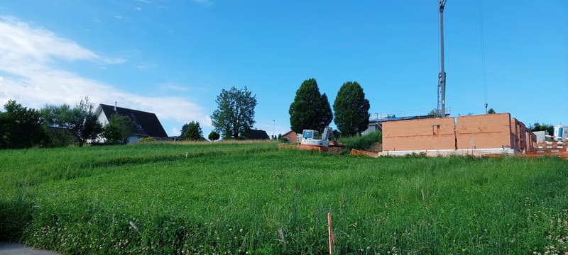 The image shows a construction site with a grassy field, some trees, and a building under construction. There is a construction vehicle visible on the site, and what appears to be a parking area or space for vehicles.