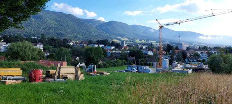 The image shows a construction site in a mountainous, forested area. There is a crane visible, along with various construction materials and equipment. The surrounding landscape features green hills, trees, and buildings in the distance, with mountains in