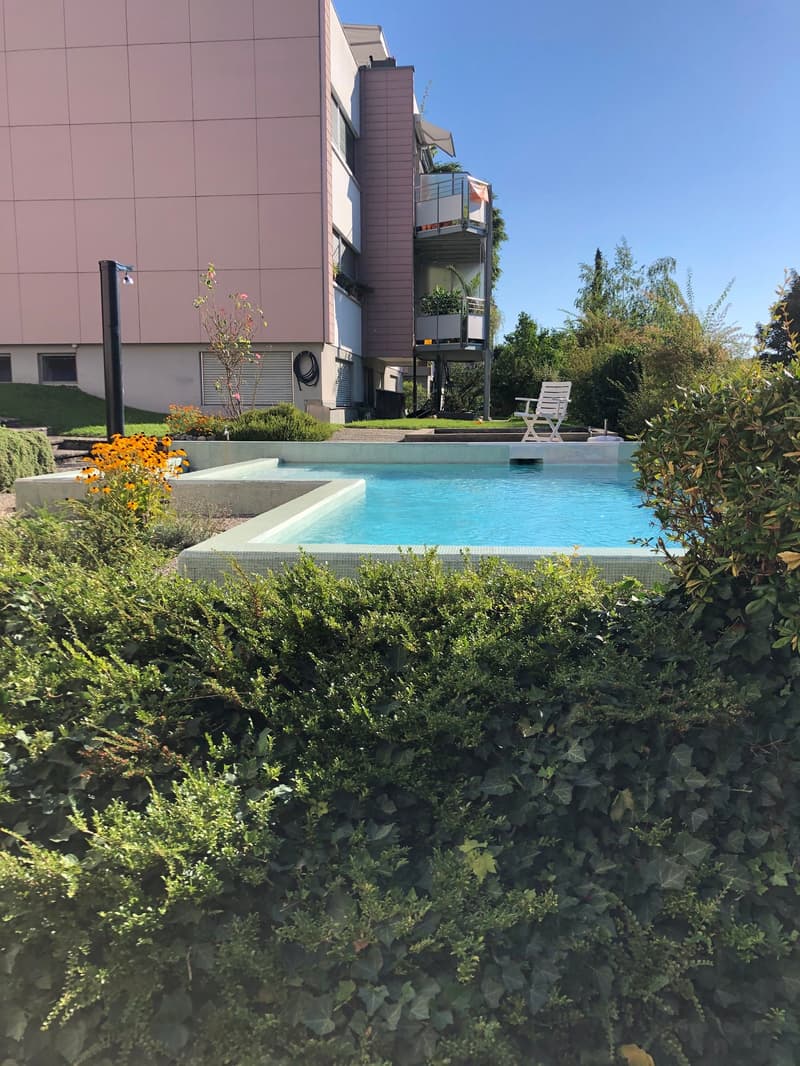 Apartment building exterior with swimming pool, beige walls, outdoor white lounge chair, shrubs, flowers
