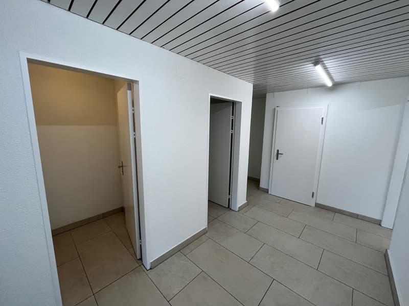 Empty room with white walls and tiled floor, two open doors, one of which reveals an adjacent room, ceiling lights
