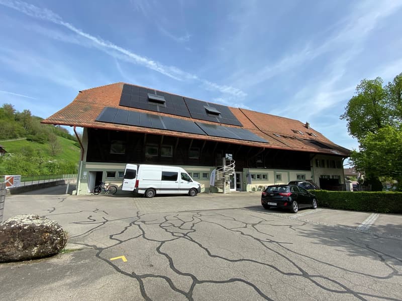 A two-story commercial building with a red tile roof and solar panels installed on the roof. The building has large windows and a spiral staircase on the exterior. There are two vehicles parked in the paved parking area in front of the building.