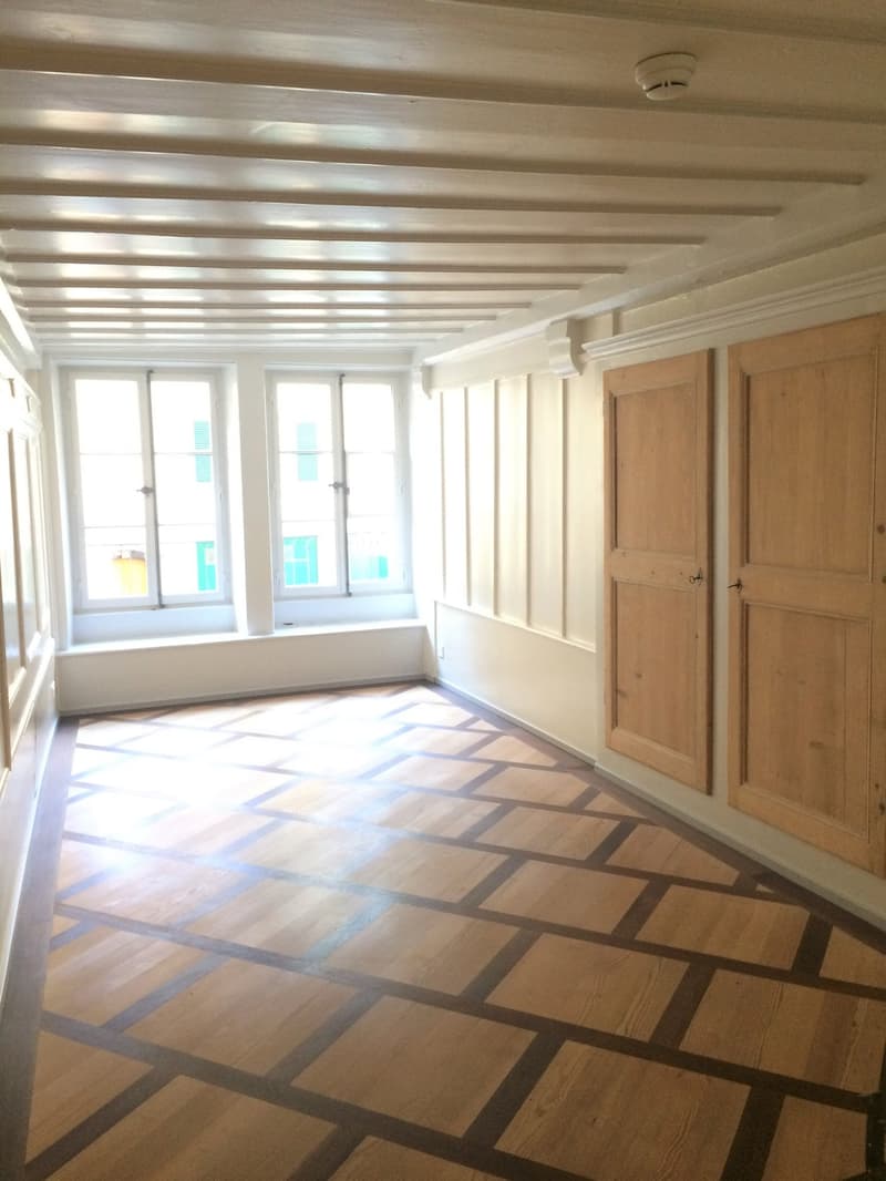 Wooden floor, white ceiling, white wall panels, wooden doors, three windows, window bench, and outside view