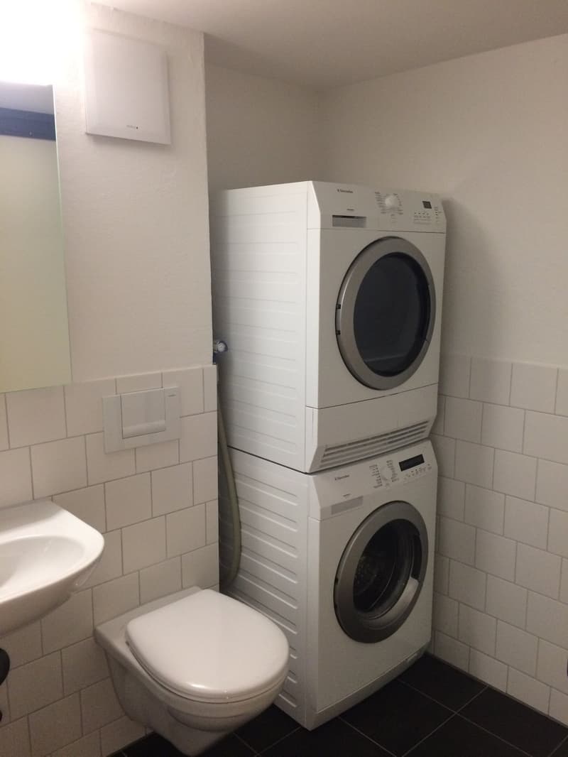 small bathroom with white tiles, toilet, sink, stackable washing machine and dryer