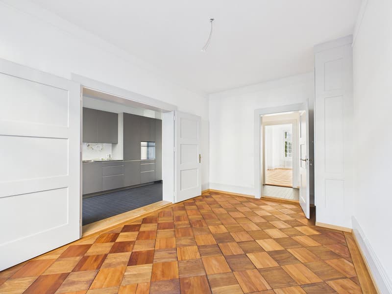 Empty room, wooden floor, white walls, open doors, kitchen in the background