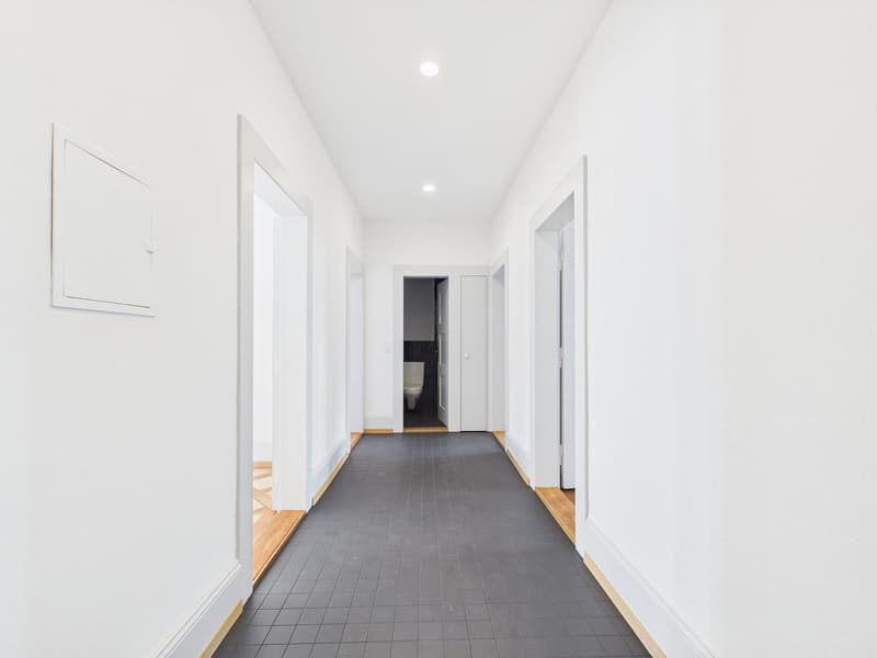 Empty hallway with white walls, black tiled floor, wooden baseboards, and recessed lighting.