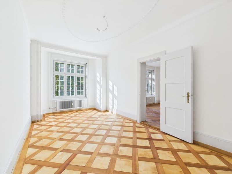 Empty room with wooden parquet flooring, white walls, large windows, radiator, open door