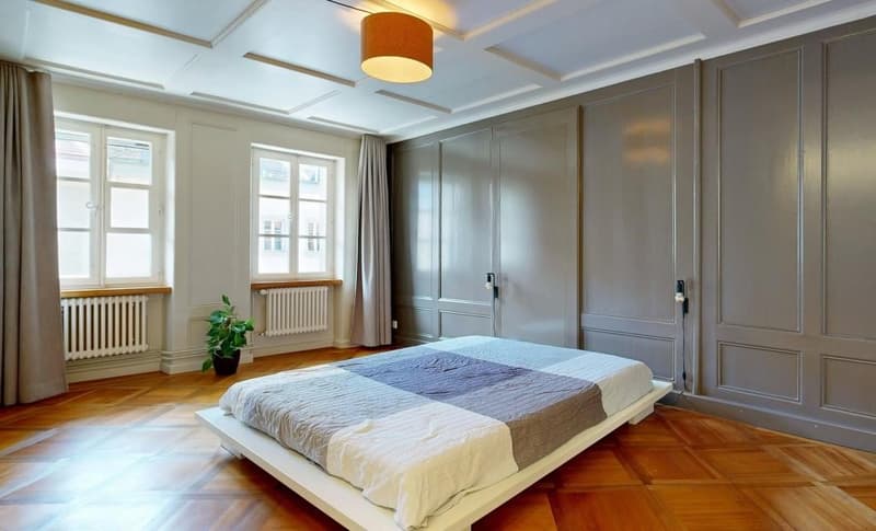 Bedroom with wooden floor, bed, potted plant, radiators, windows, gray walls, ceiling lamp