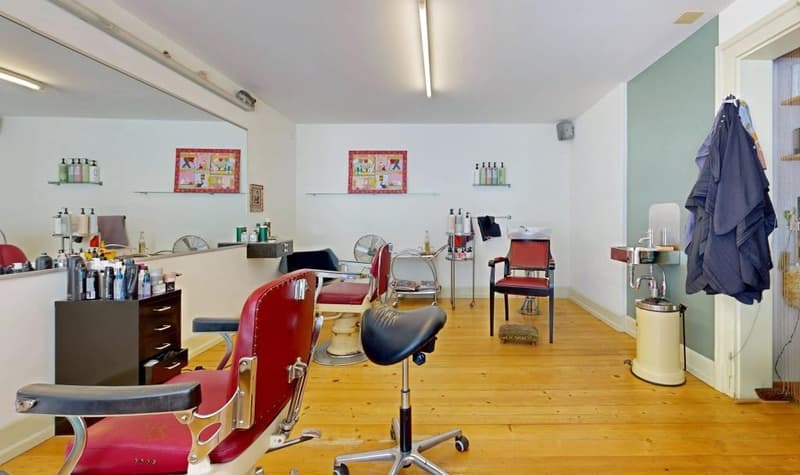 A room with a wooden floor, a large mirror, a cabinet, a few chairs, a desk, and a trash can.