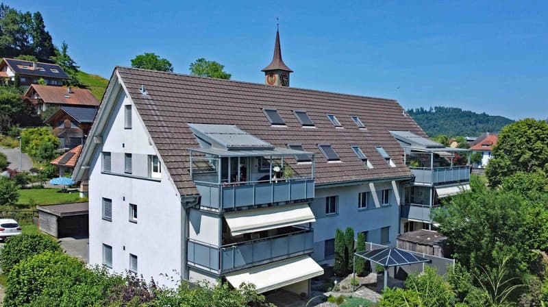 A two-story building with brown roof and white walls. It features multiple balconies and windows. The area around the building is surrounded by greenery, and there is a clock tower on top of the building.