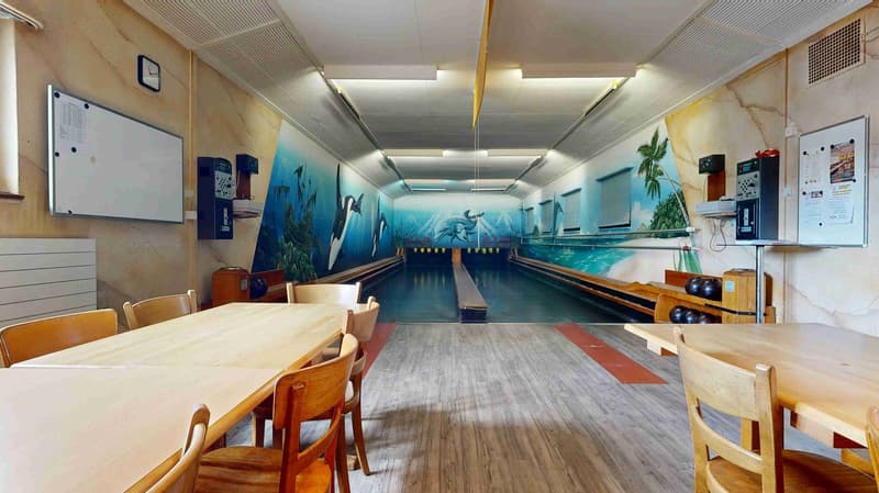 Dining area with wooden tables and chairs, bowling alley with lanes and ball return, walls with murals, whiteboards, and a clock