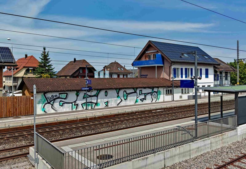 Train station with graffiti on the wall, solar panels on the roof, houses in the background, and a platform with a shelter