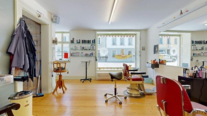 Barber shop with wooden floor, white walls, shelves with products, barber chairs, mirror, radiator, trash bin, and a door.