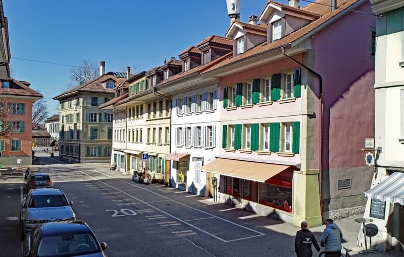 A row of buildings with green shutters, a street with parked cars, and people walking on the sidewalk.