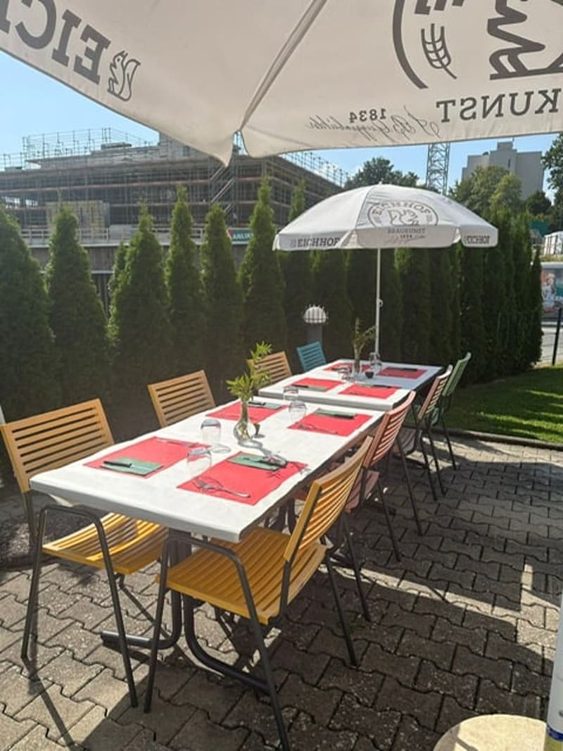 tables with red and white tablecloths, wooden chairs, umbrella, green grass, bushes, building under construction