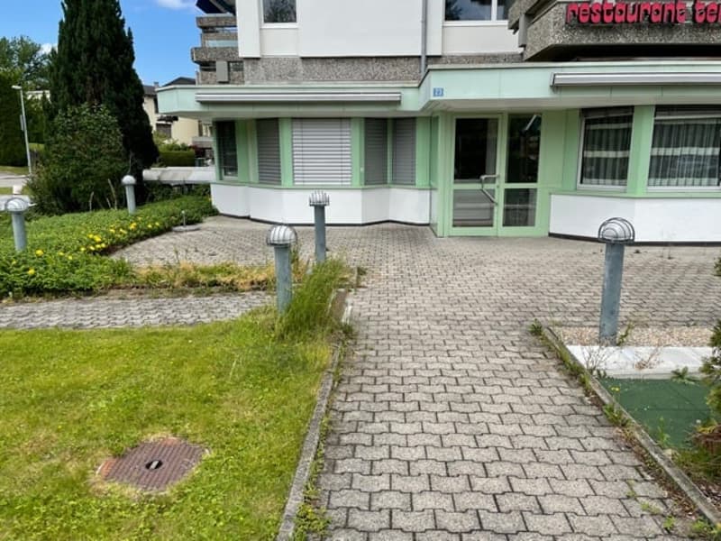 Building facade with green window frames and a walkway in front, with plants and grass on the sides.