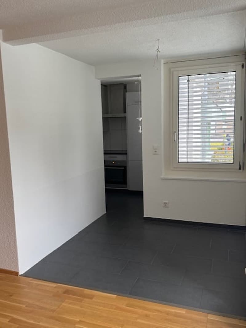 Empty room, white walls, black tile flooring, wooden floor, window with blinds, doorway to another room, power outlet, kitchen area