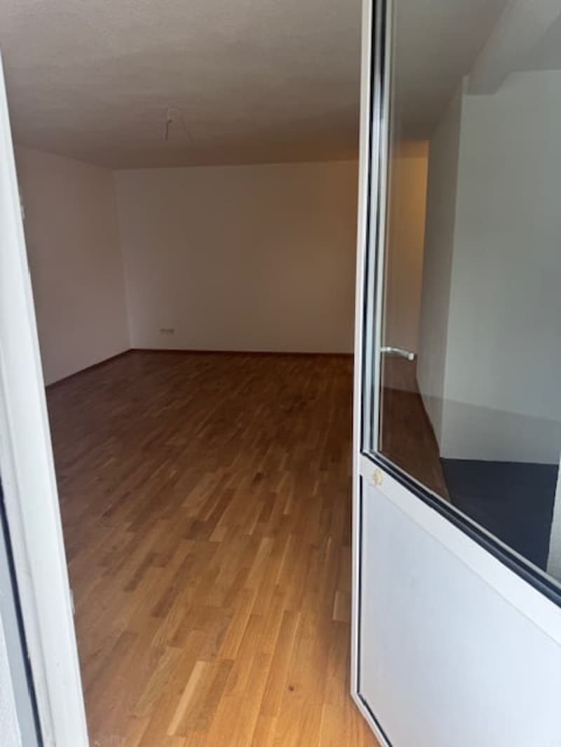 Empty room with hardwood floors, white walls, open door leading to hallway, a small object is placed on the floor on the left side of the image.
