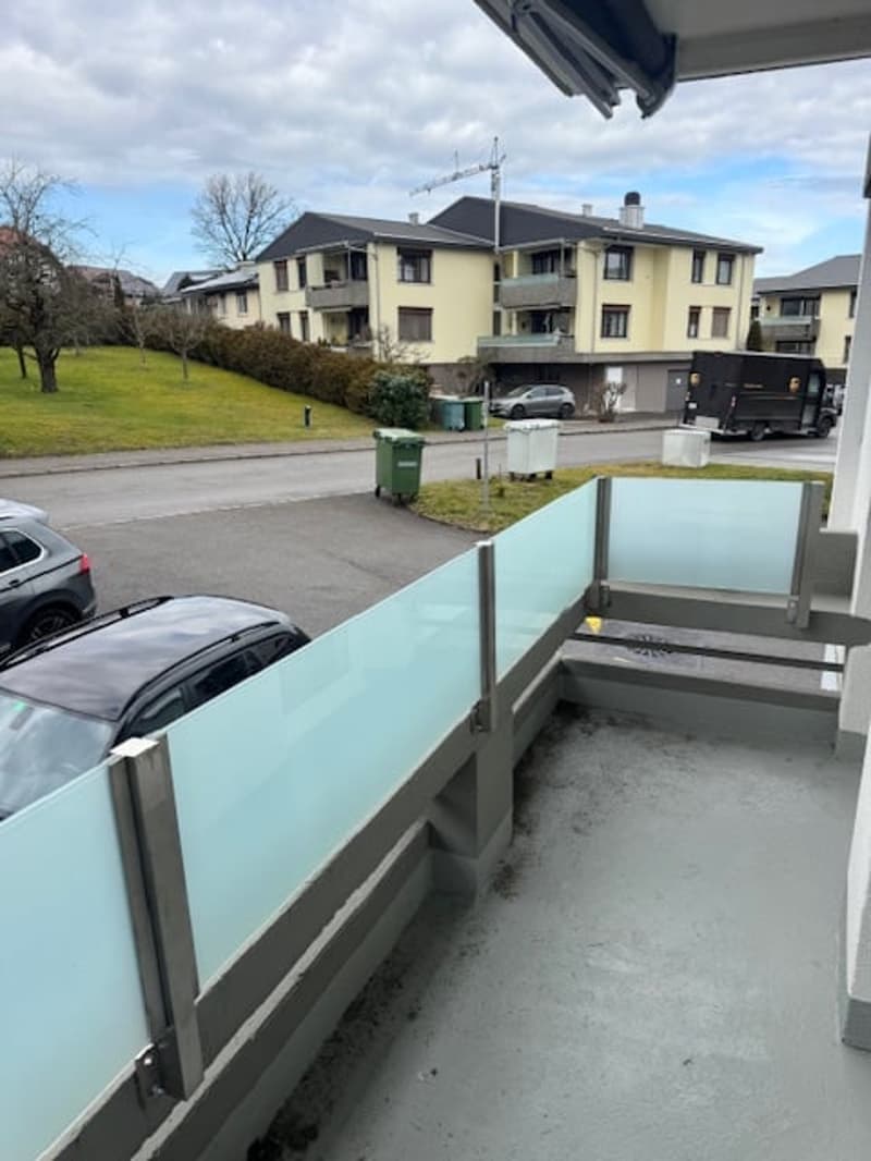 balcony with glass railing, concrete floor, cars parked on the street, grass and buildings in the distance