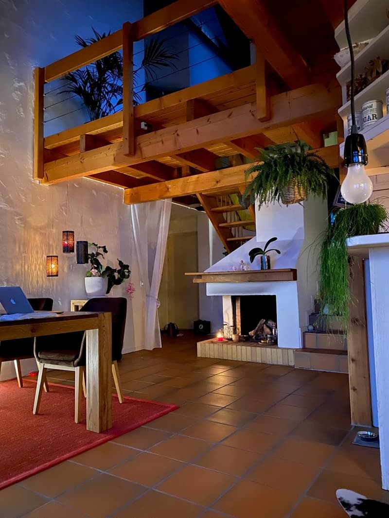 Living area with fireplace, dining table, wooden chairs, red rug, laptop, potted plants, wooden balcony with railing, ceiling lamps, white walls, staircase.