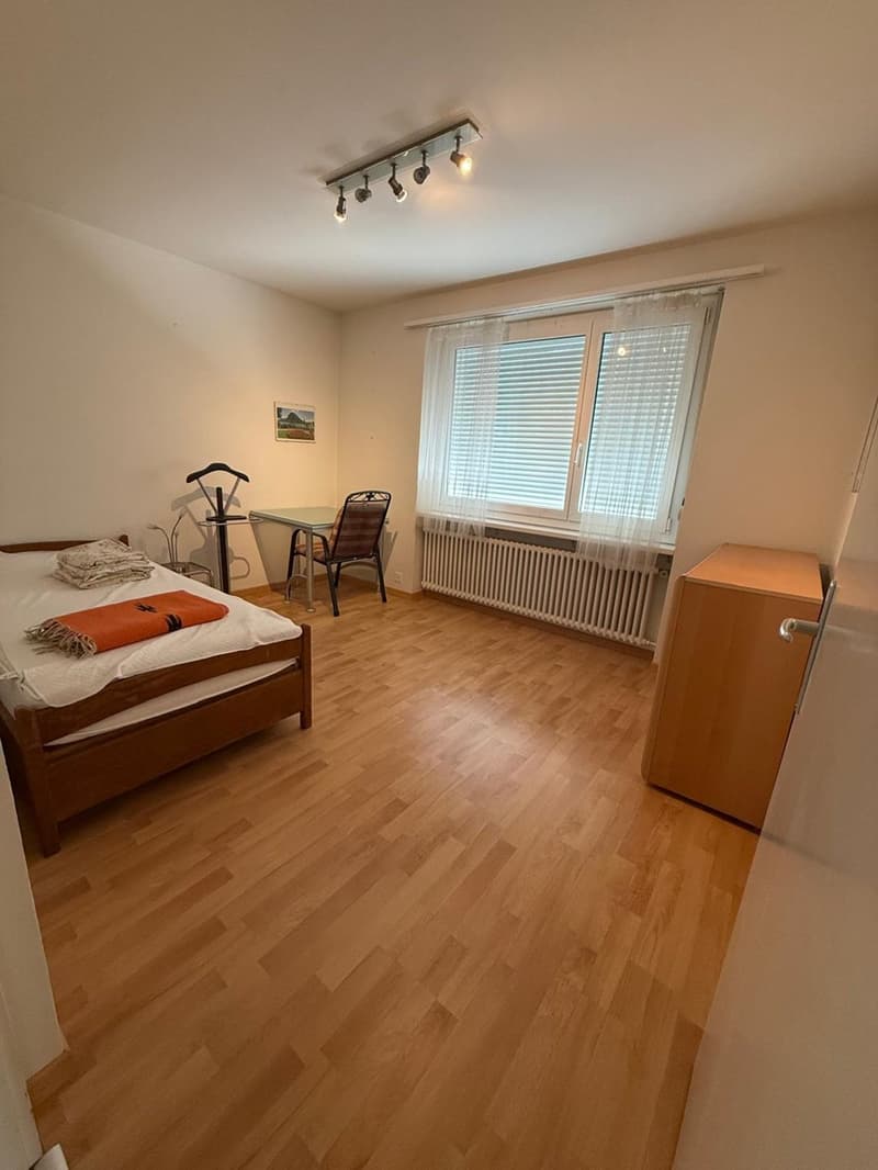 Bedroom with wooden bed, desk, chair, radiator, window with curtains, and wooden cabinet.