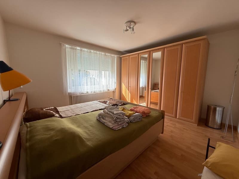 room contains two single beds, desk with lamp, wardrobe, waste bin, large window with curtain, ceiling light