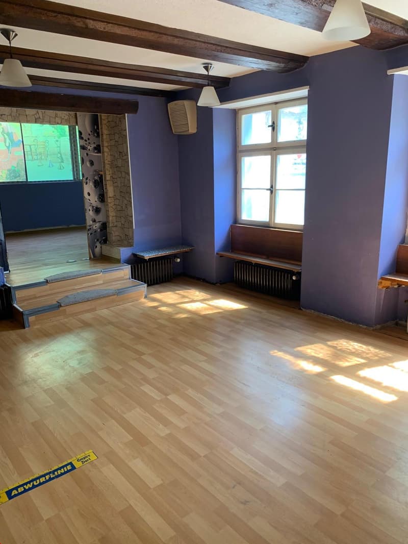 Large living room with hardwood floors, exposed wooden beams on the ceiling, and a fireplace. The room has large windows that let in natural light. There are built-in benches along the walls and a chalkboard or bulletin board on the wall.