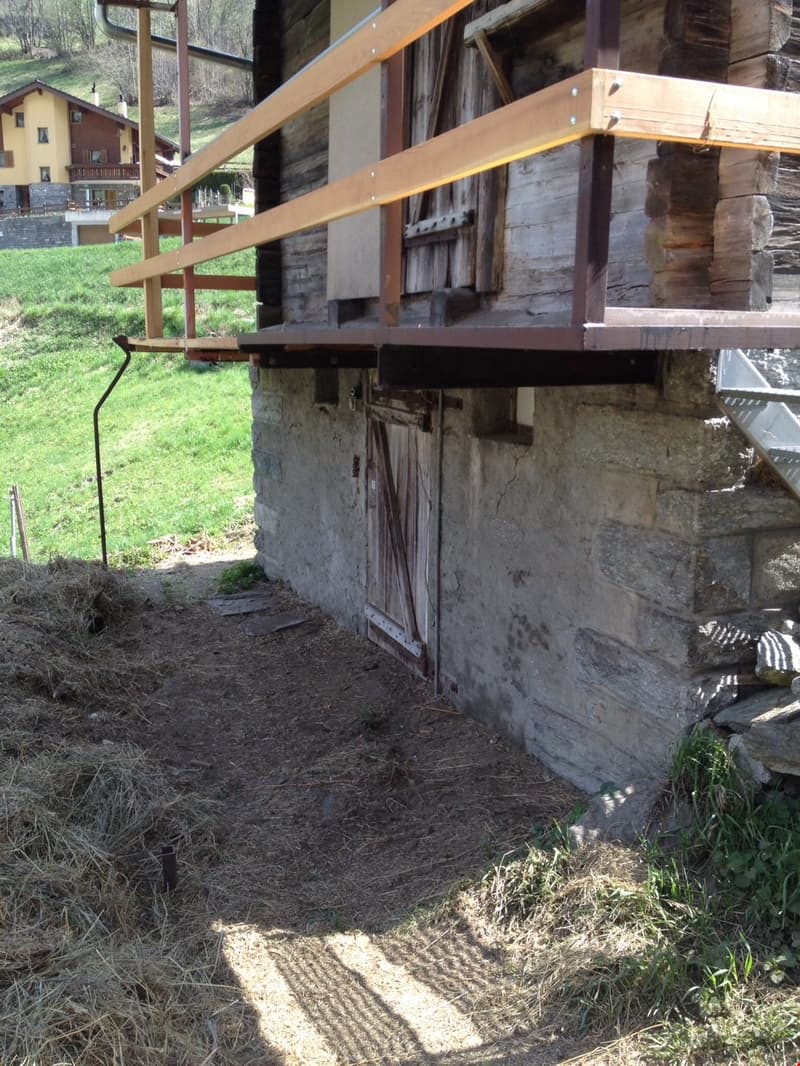 A rustic stone building with a wooden overhang and a dirt path leading to it. The building appears to be situated in a grassy, natural setting with some other structures visible in the background.