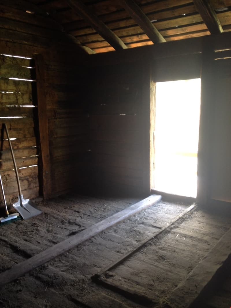 The image shows the interior of an attic space. The walls are made of wooden boards, and the floor is covered in dirt and debris. There is a bright opening, likely a window or doorway, that is letting in natural light from the outside.