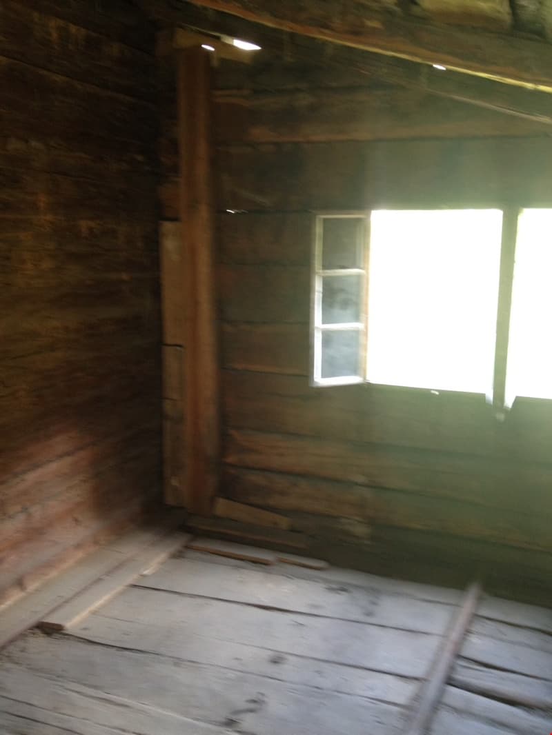 The image shows the interior of a wooden cabin or shed. The walls are made of wooden planks, and there is a small window that lets in natural light. The floor appears to be made of wooden boards as well. The overall atmosphere is dark and rustic, with a s