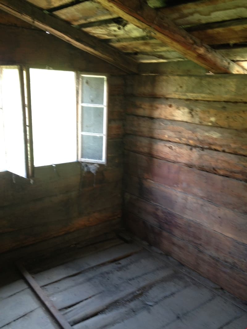 The image shows the interior of an old, rustic cabin or shed. The walls are made of weathered, wooden logs, and the floor appears to be made of wooden planks. There is a small window that allows natural light to enter the space. The overall atmosphere is