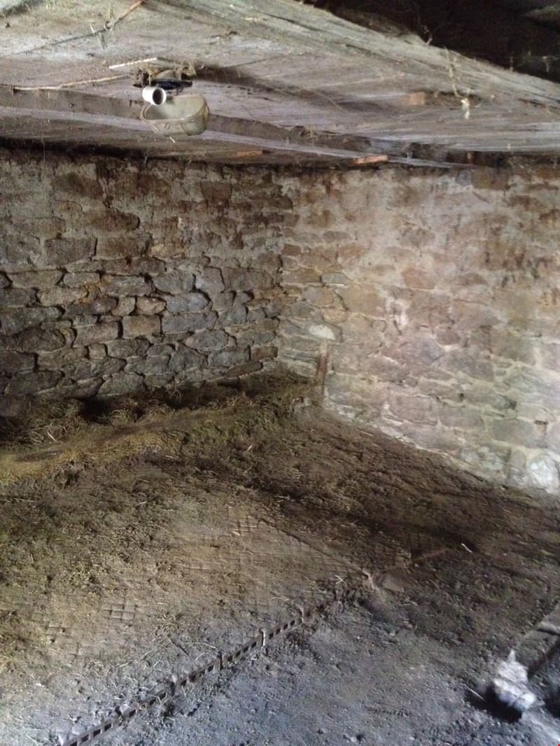 The image shows the interior of a basement or cellar. The walls are made of rough, unfinished stone or concrete, and the floor appears to be dirt or gravel. There is some debris and clutter visible on the floor, and an old, rusted pipe or vent can be seen