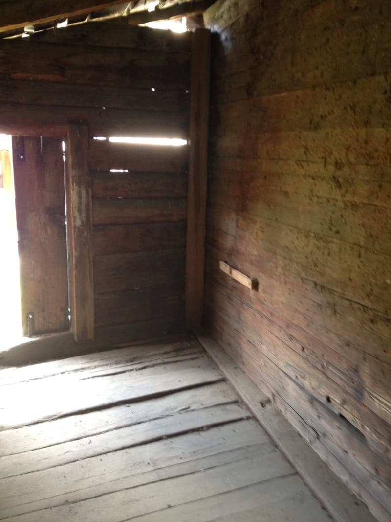 The image shows the interior of an old, rustic building. The walls are made of rough, unfinished wood, and the floor is also wooden. There is a doorway visible, leading to another room or area. The lighting is dim, with some natural light coming in throug