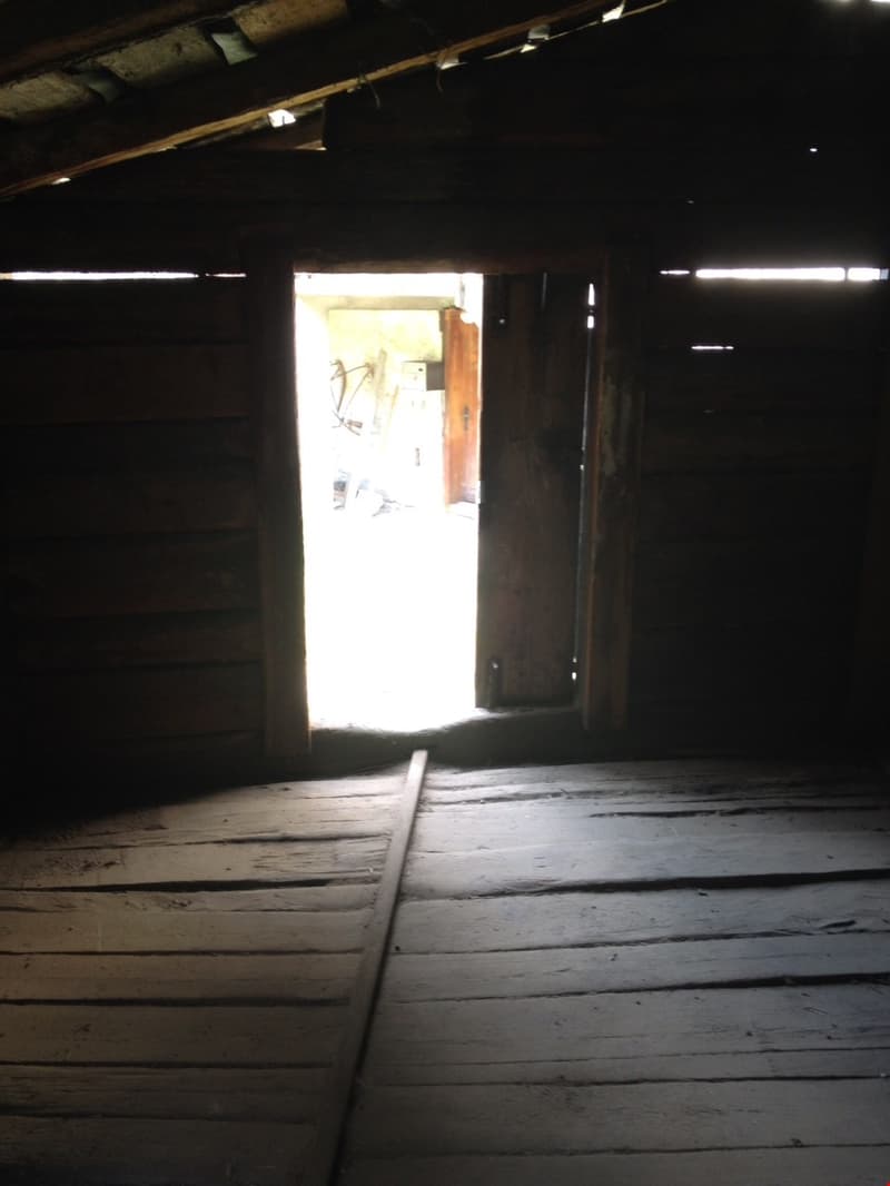 Dark, wooden attic space with a doorway leading to a brighter area. The floor is made of wooden planks and the ceiling has exposed beams.